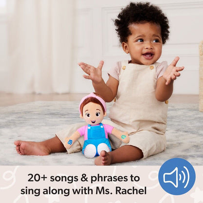 A happy baby with curly hair and a Ms. Rachel plush doll sit on a rug, the baby smiles, hands raised, wearing a beige romper
