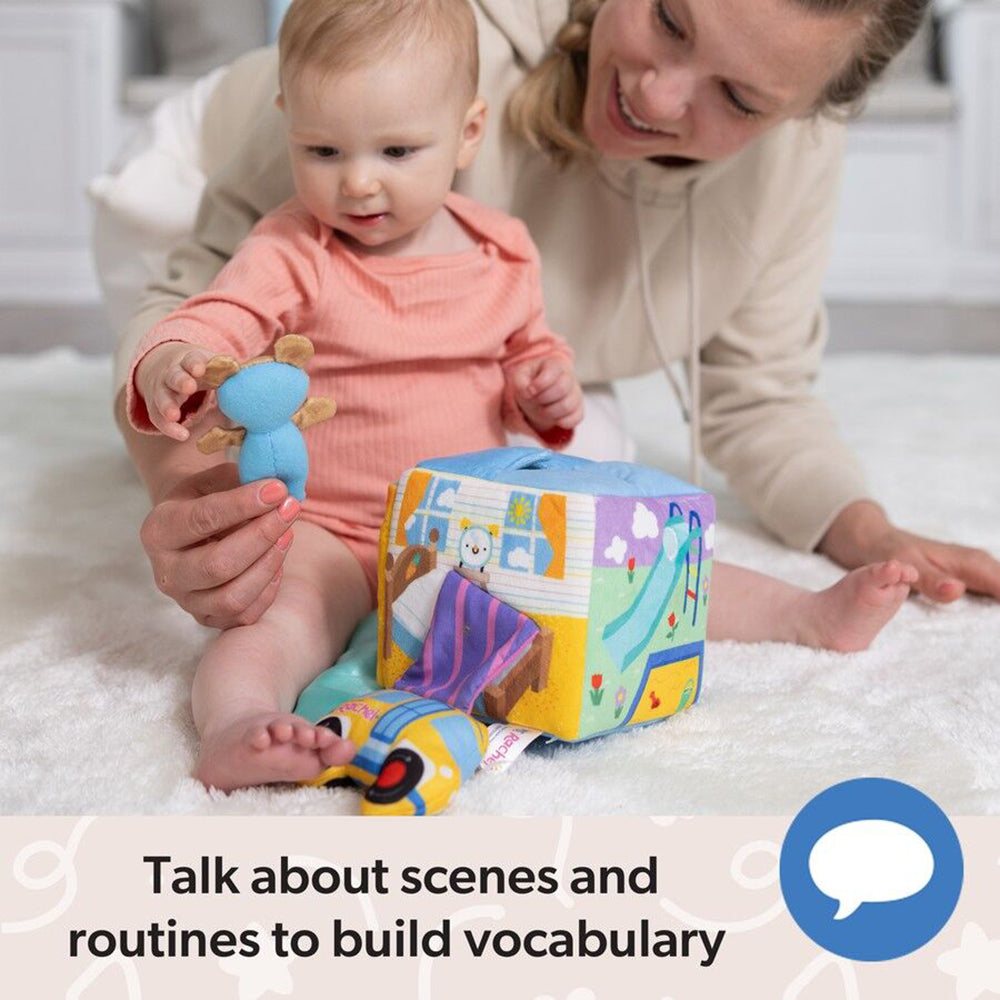 Baby in pink holds a blue toy, playing with a colorful activity cube featuring bedroom and playground scenes, watched by a smiling adult