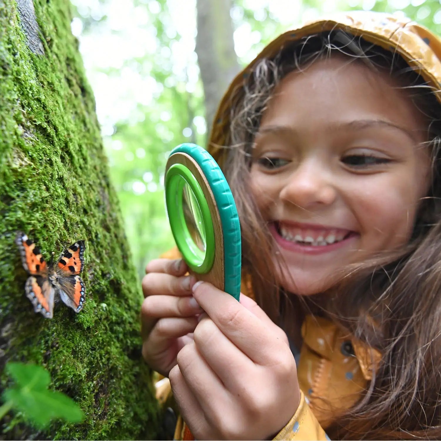 Smiling girl using a magnifying glass to study a butterfly on a mossy tree