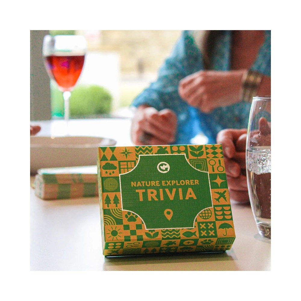 A green &quot;Nature Explorer Trivia&quot; game box sits on a table, with people&#39;s hands and drinks blurred in the background