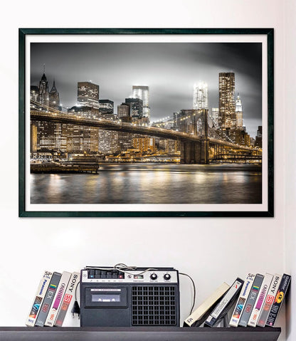 Framed black and white photo of Brooklyn Bridge & illuminated NYC skyline at night, golden lights reflecting on water