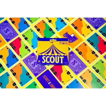 A SCOUT card game box, with its blue and yellow circus tent design, is surrounded by colorful, numbered cards fanned out on a yellow surface