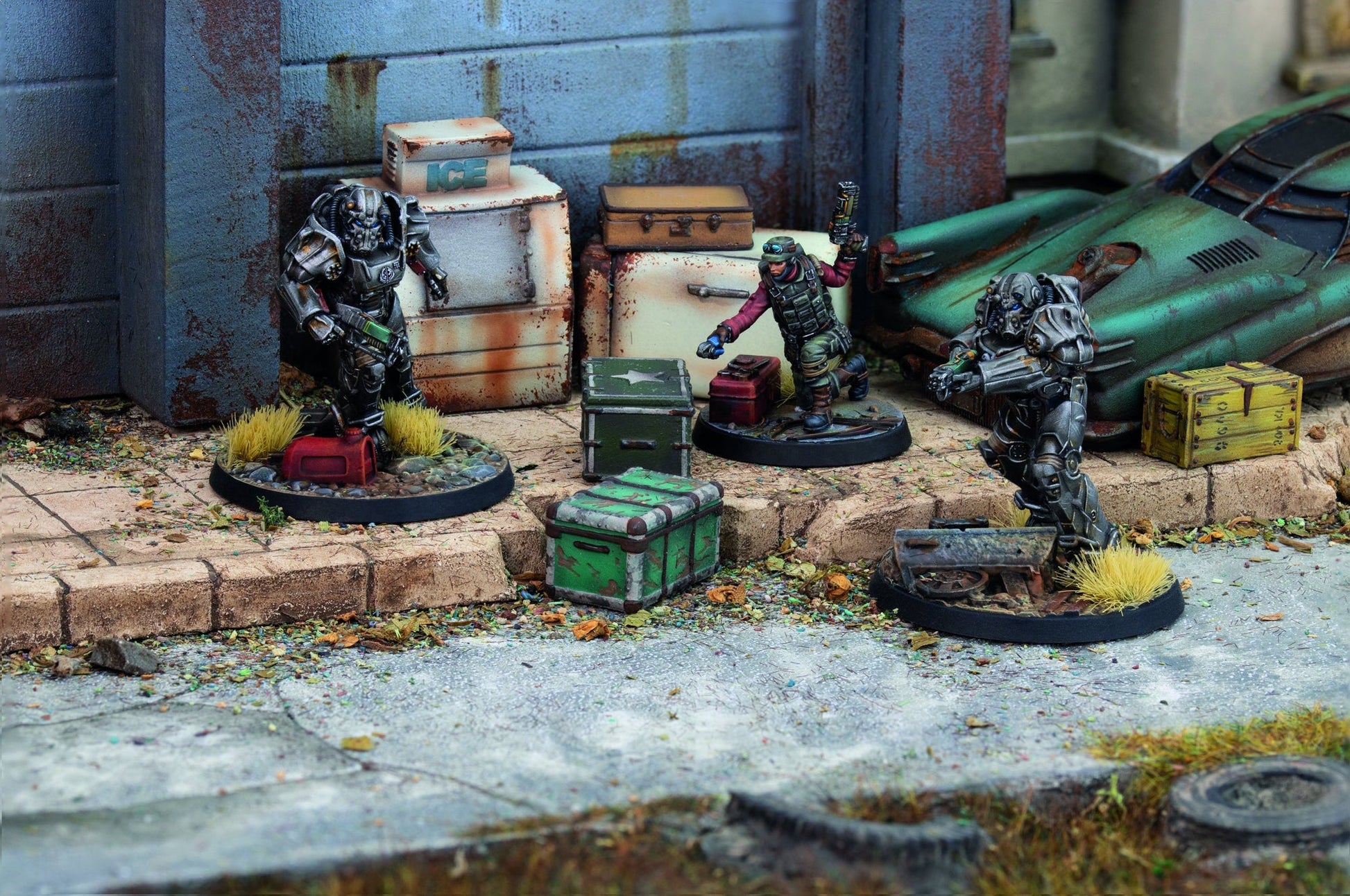 Three sci-fi miniatures: two power-armored, one tactical human, scavenging amidst debris, crates, and a rusted car