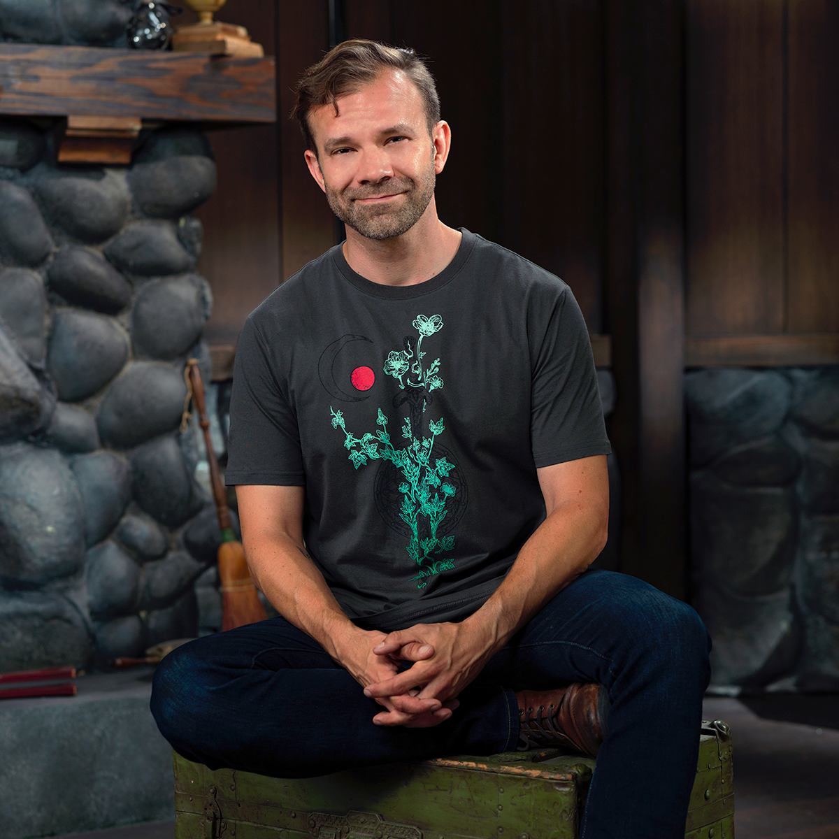 A smiling bearded man sits cross-legged on a green trunk in a rustic room, wearing a grey t-shirt with a green plant & red moon design