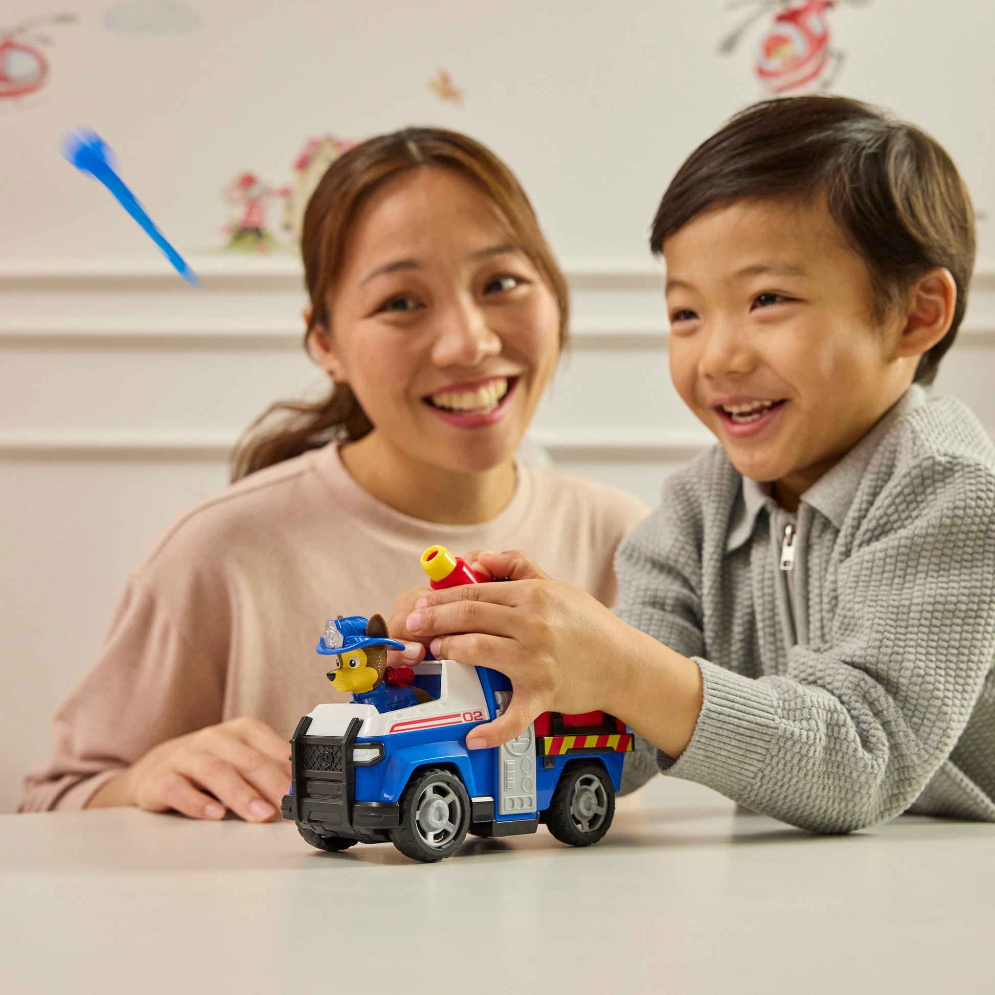 Smiling boy plays with a blue Paw Patrol police truck toy featuring Chase, a happy woman looks on, and a blue dart flies in the background