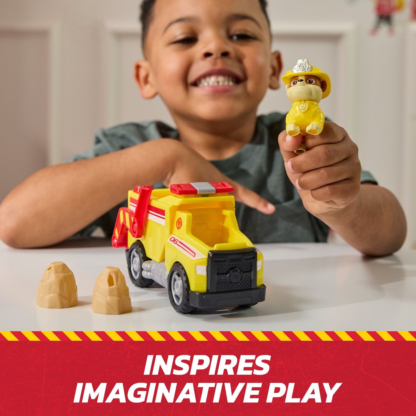 Smiling child holding a yellow Paw Patrol Rubble toy, playing with his matching yellow construction truck and two tan rocks on a table