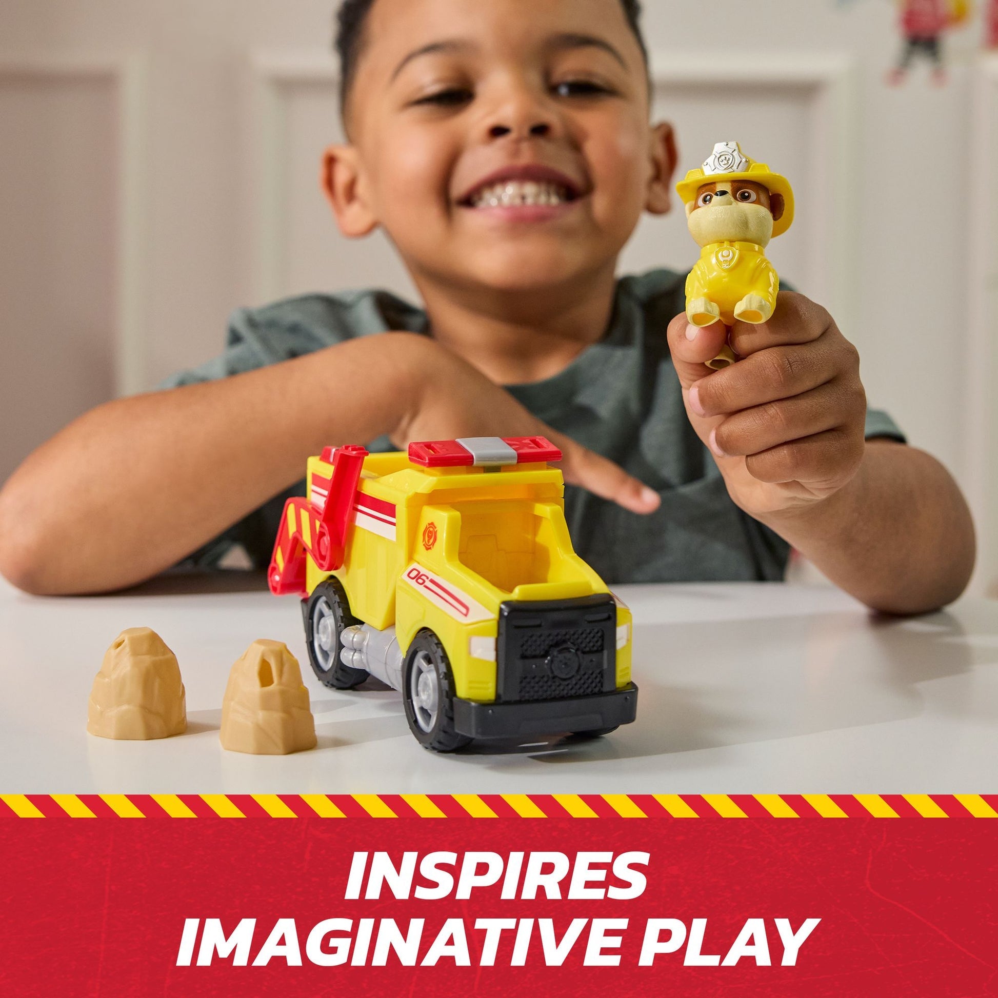 Smiling child holding a yellow Paw Patrol Rubble toy, playing with his matching yellow construction truck and two tan rocks on a table