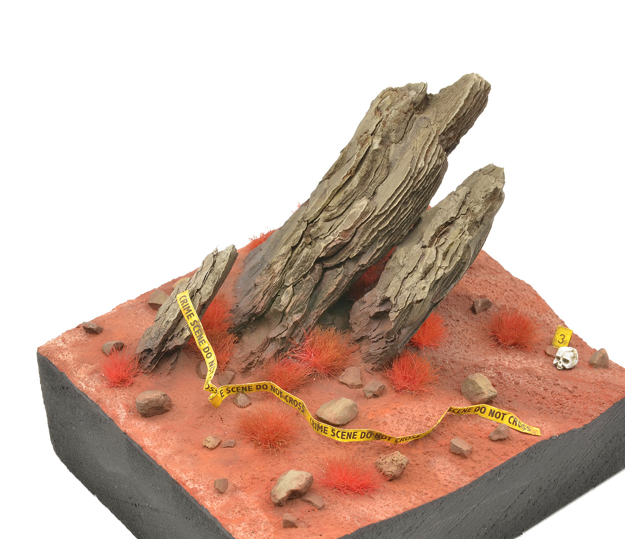 A miniature diorama showcases bright red AK Interactive 4mm wargame tufts scattered across red earth with large rocks, yellow crime scene tape, and a tiny skull