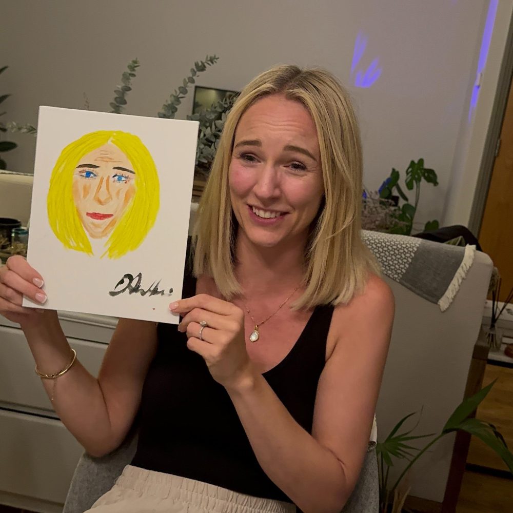 A smiling blonde woman holds up a simple painting of a blonde face with blue eyes and yellow hair