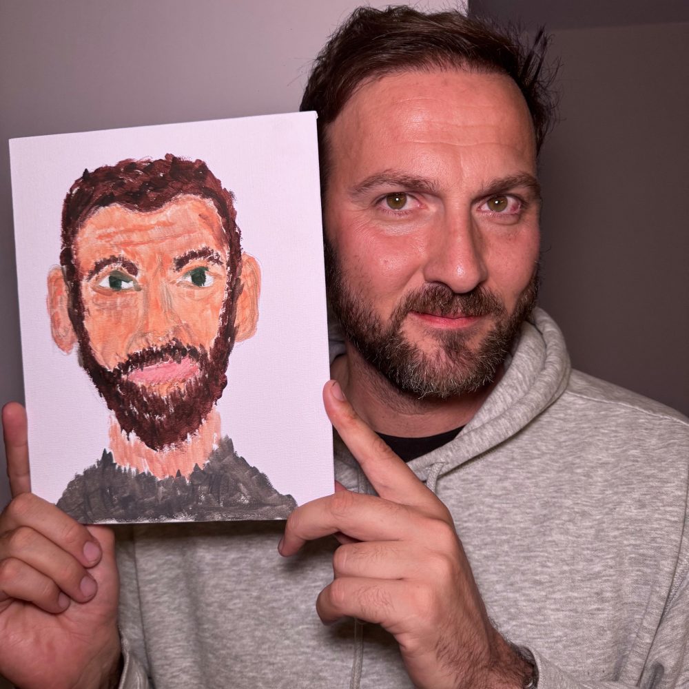 Smiling man with a beard holds up a colorful, stylized painting of himself