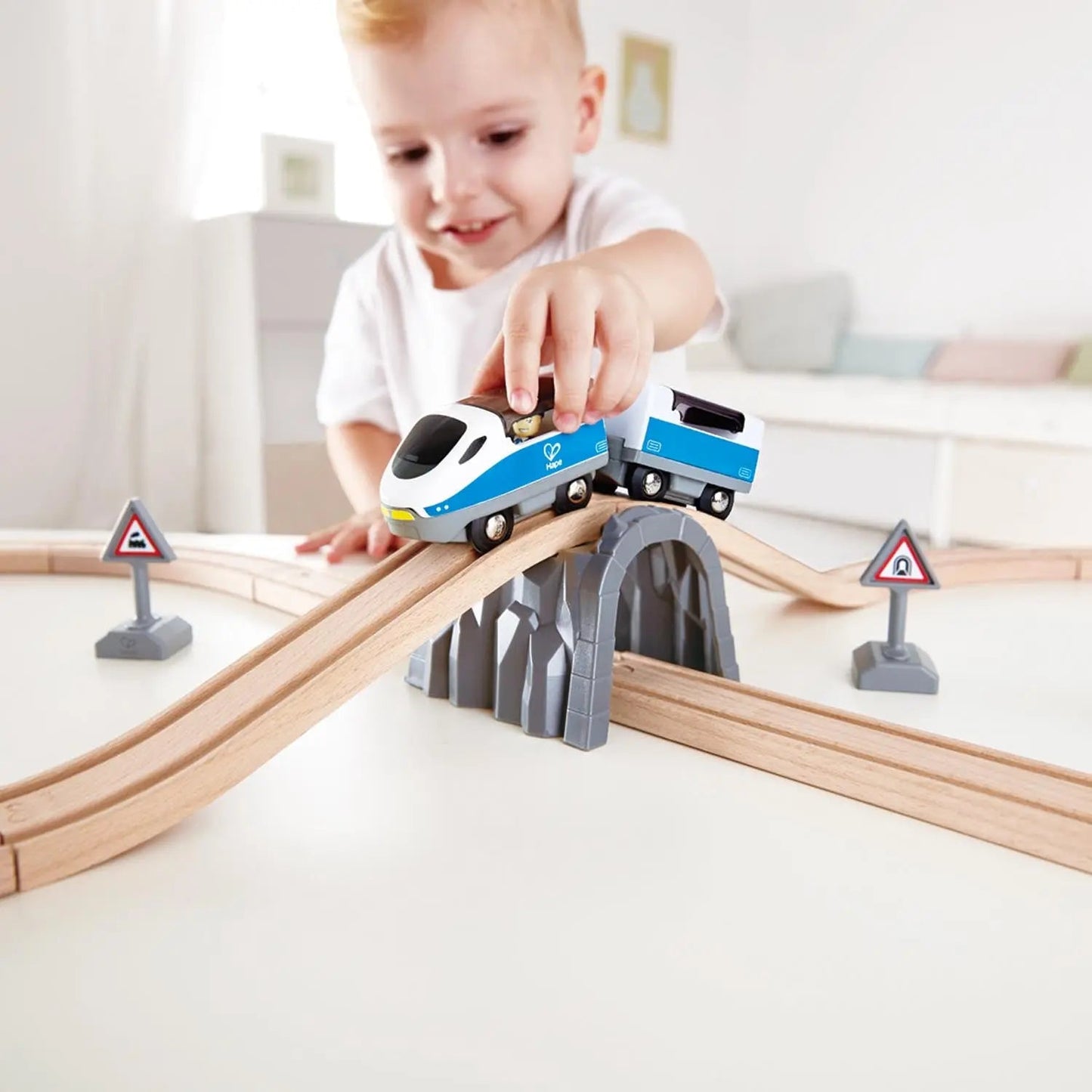 Young child pushing a blue toy train along a wooden track set with a tunnel