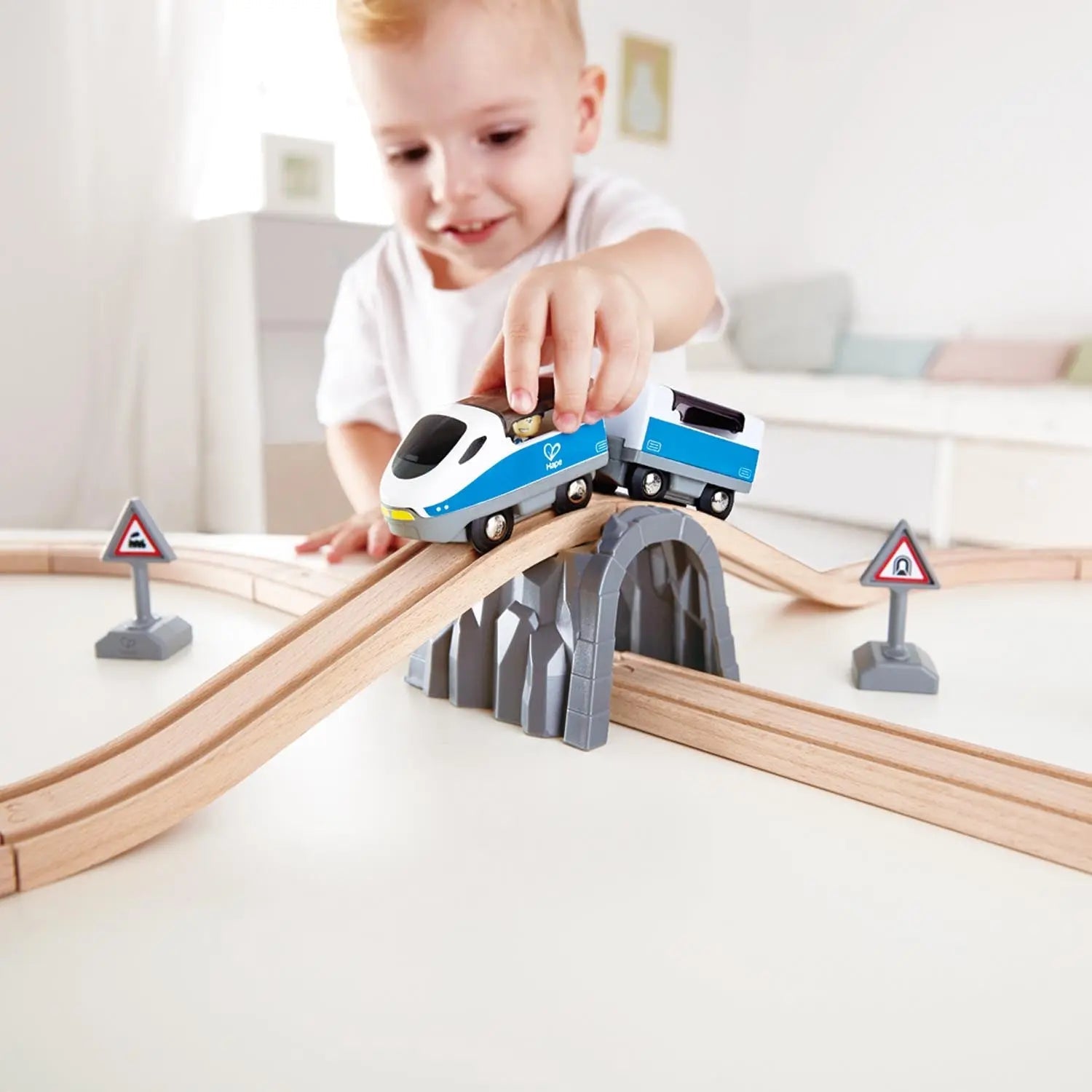 Young child pushing a blue toy train along a wooden track set with a tunnel