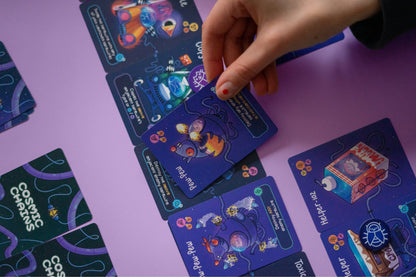 A hand holds a dark blue space-themed game card over other "Cosmic Chains" cards on a purple surface