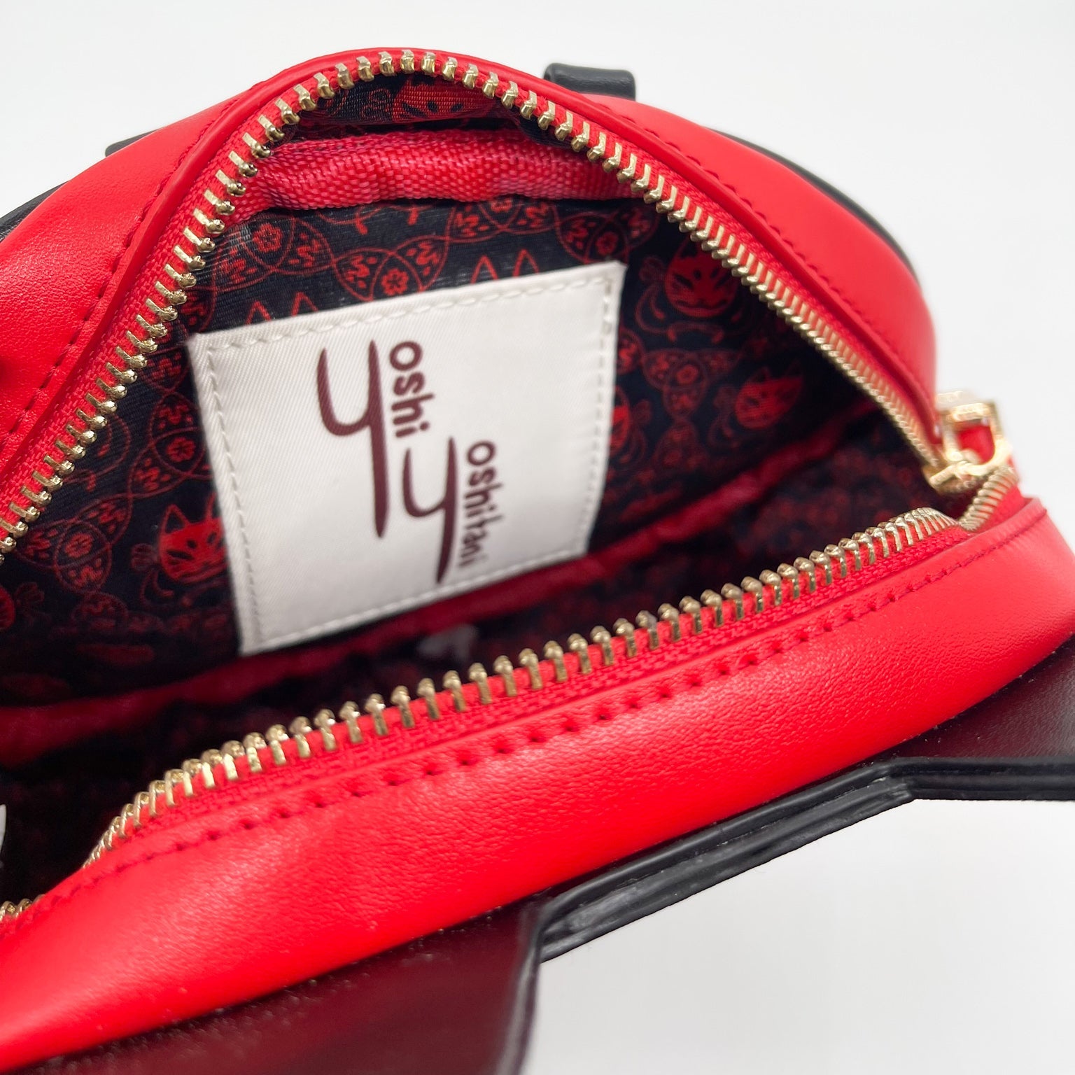 Open red leather bag with gold zipper, revealing black lining with red fox mask pattern and a "Yoshi Oshitai" label.