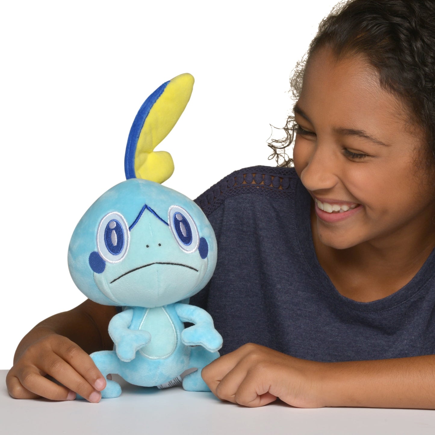 Happy girl smiles down at a blue Sobble plush toy, hands resting near it