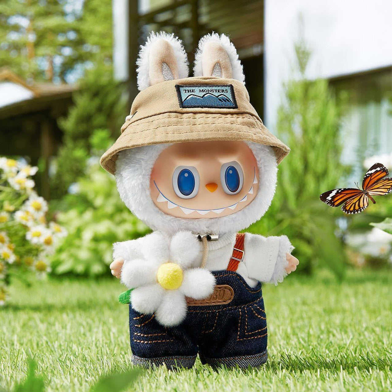 Furry white monster toy in a bunny-eared bucket hat and overalls holds a flower. A butterfly flies nearby in a sunny garden