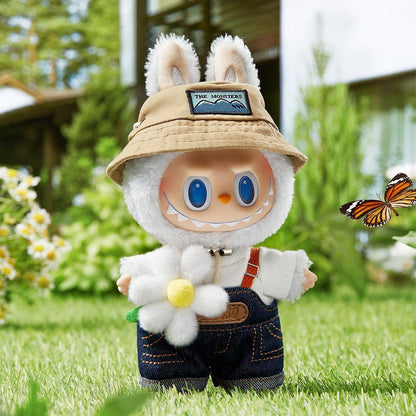 Furry white monster toy in a bunny-eared bucket hat and overalls holds a flower. A butterfly flies nearby in a sunny garden