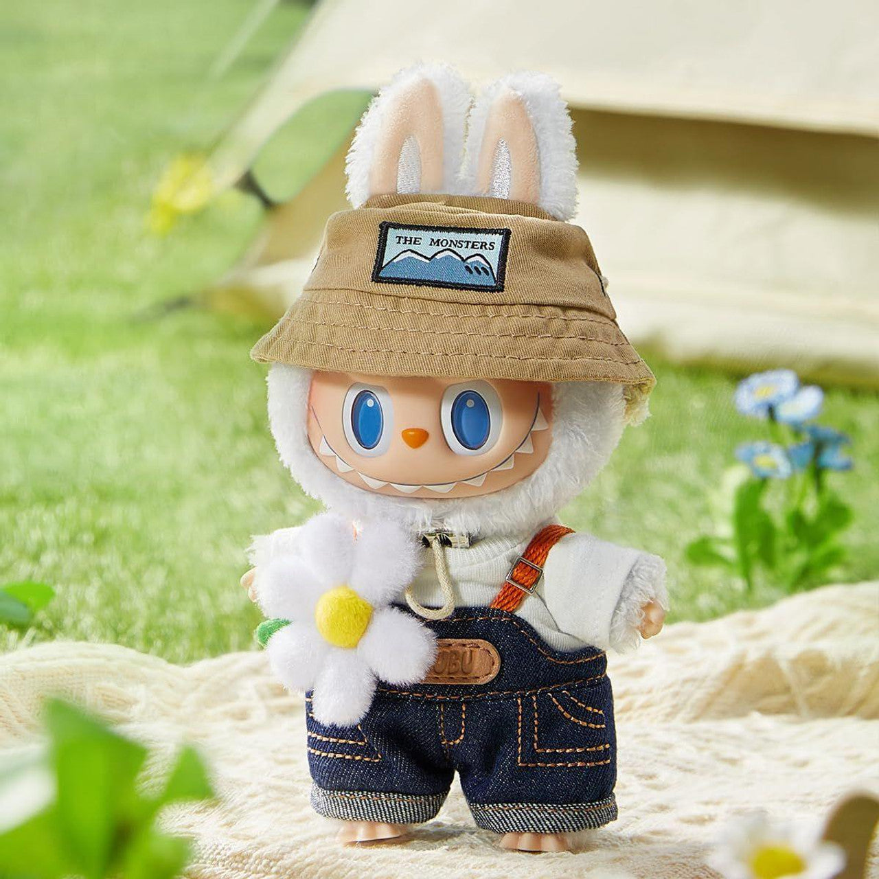 Fluffy white toy monster with bunny ears, blue eyes, wearing a bucket hat, denim overalls, holding a daisy. Outdoors on a picnic blanket