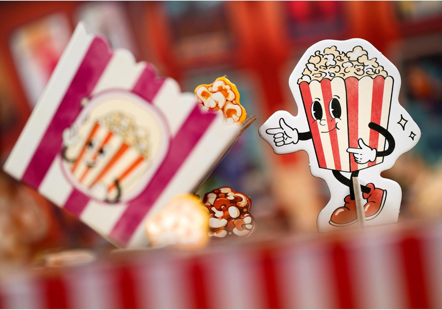 A red and white striped cartoon popcorn box character points, with a blurred purple popcorn box and small popcorn decor nearby