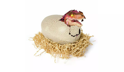 Hatching raptor toy with glowing yellow eyes emerging from a cracked, textured egg in a nest of shredded paper on a white background