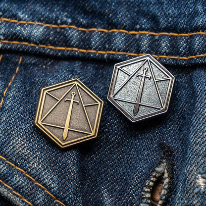 Two hexagonal Critical Role lapel pins, one gold and one silver, featuring a geometric sword emblem, on blue denim