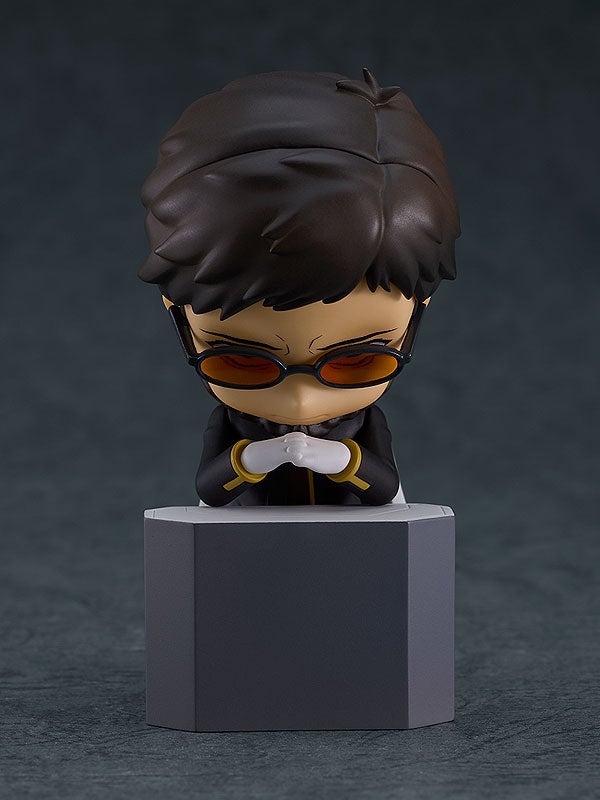 Male figurine with dark hair, orange glasses, and a black suit, hands clasped on a gray block, looking down