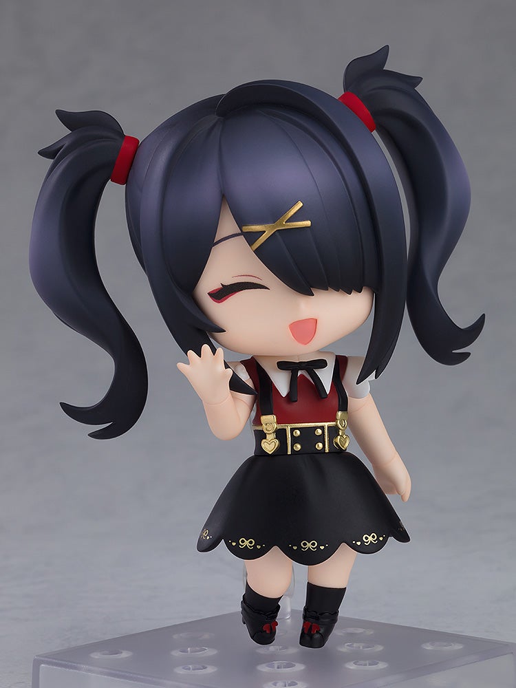 A Nendoroid figure of a dark-haired girl with pigtails, smiling and waving. She wears a red top, black skirt, and gold suspenders