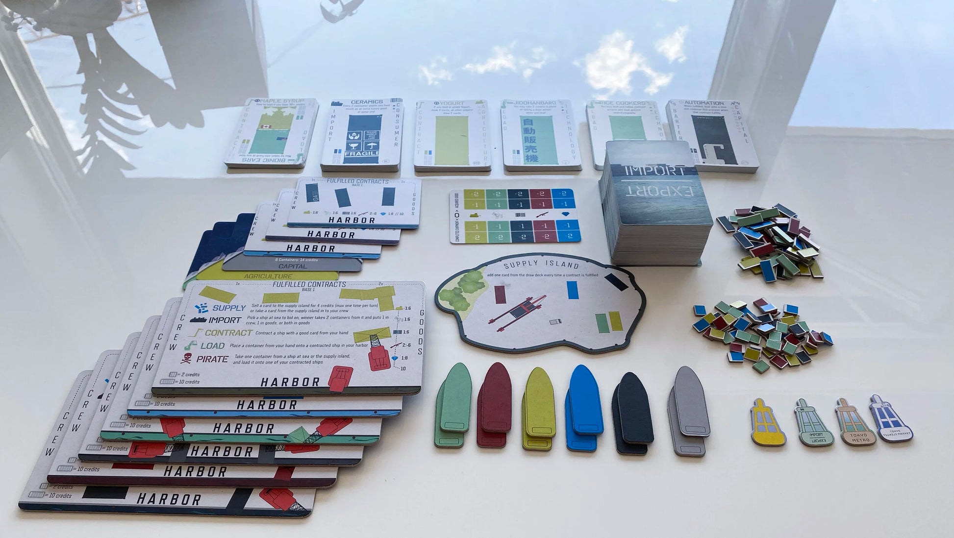 Components of the Import_Export board game are spread on a white surface, including stacks of cards, colourful ship tokens, small rectangular tokens, and the Supply Island board