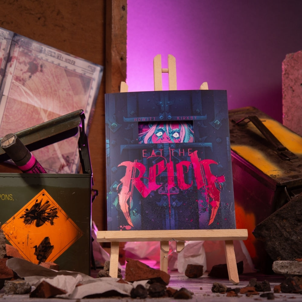 Graphic novel "EAT THE RICH" on an easel, surrounded by rubble, an ammo box, and vibrant pink backlighting