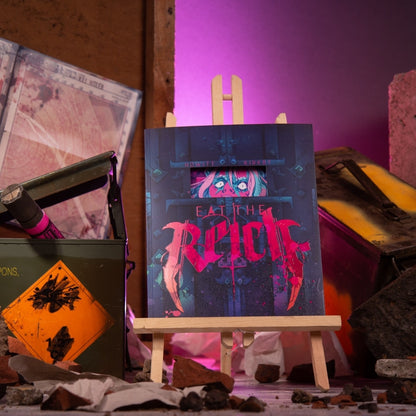 Graphic novel "EAT THE RICH" on an easel, surrounded by rubble, an ammo box, and vibrant pink backlighting