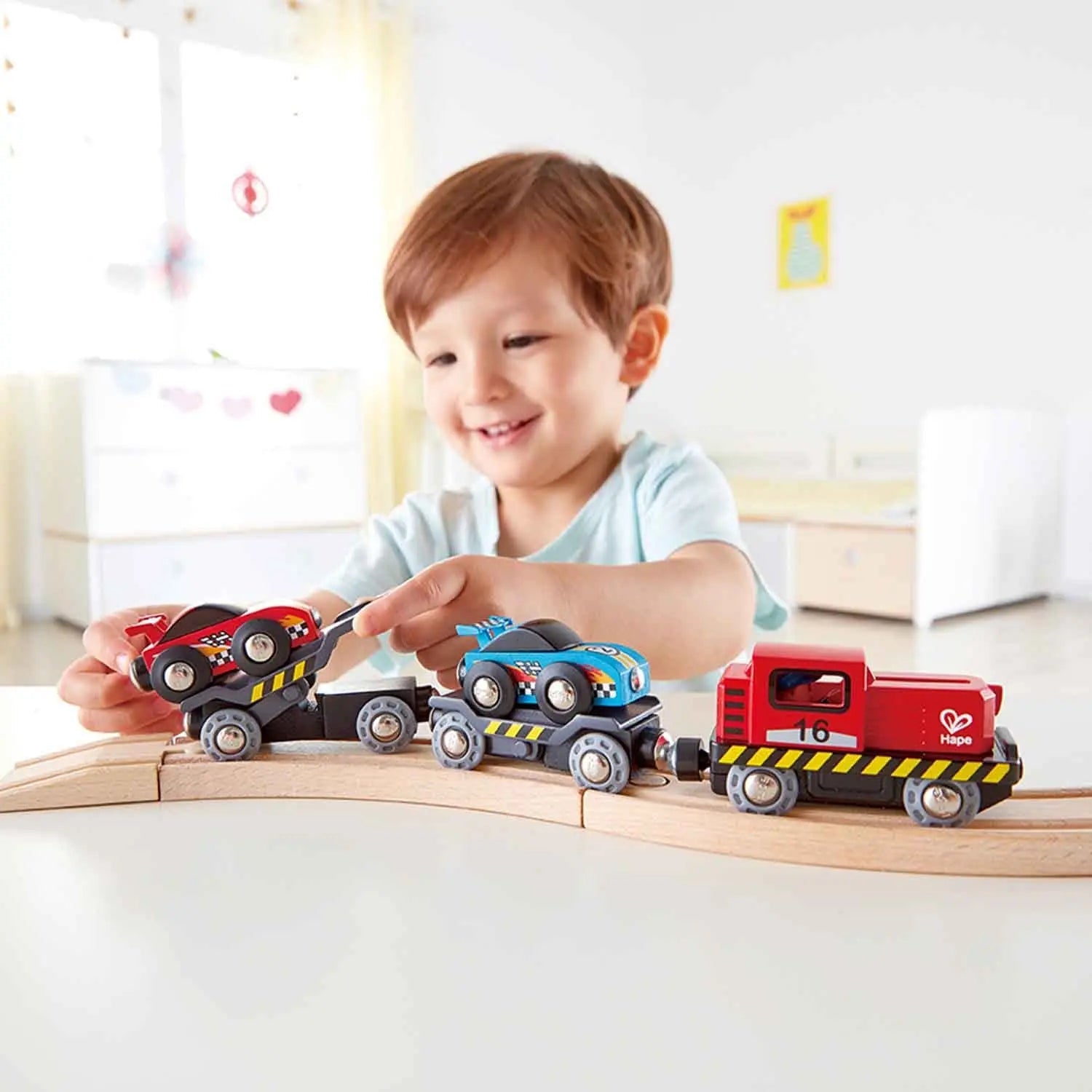 Smiling toddler interacting with a wooden train set transporting race cars