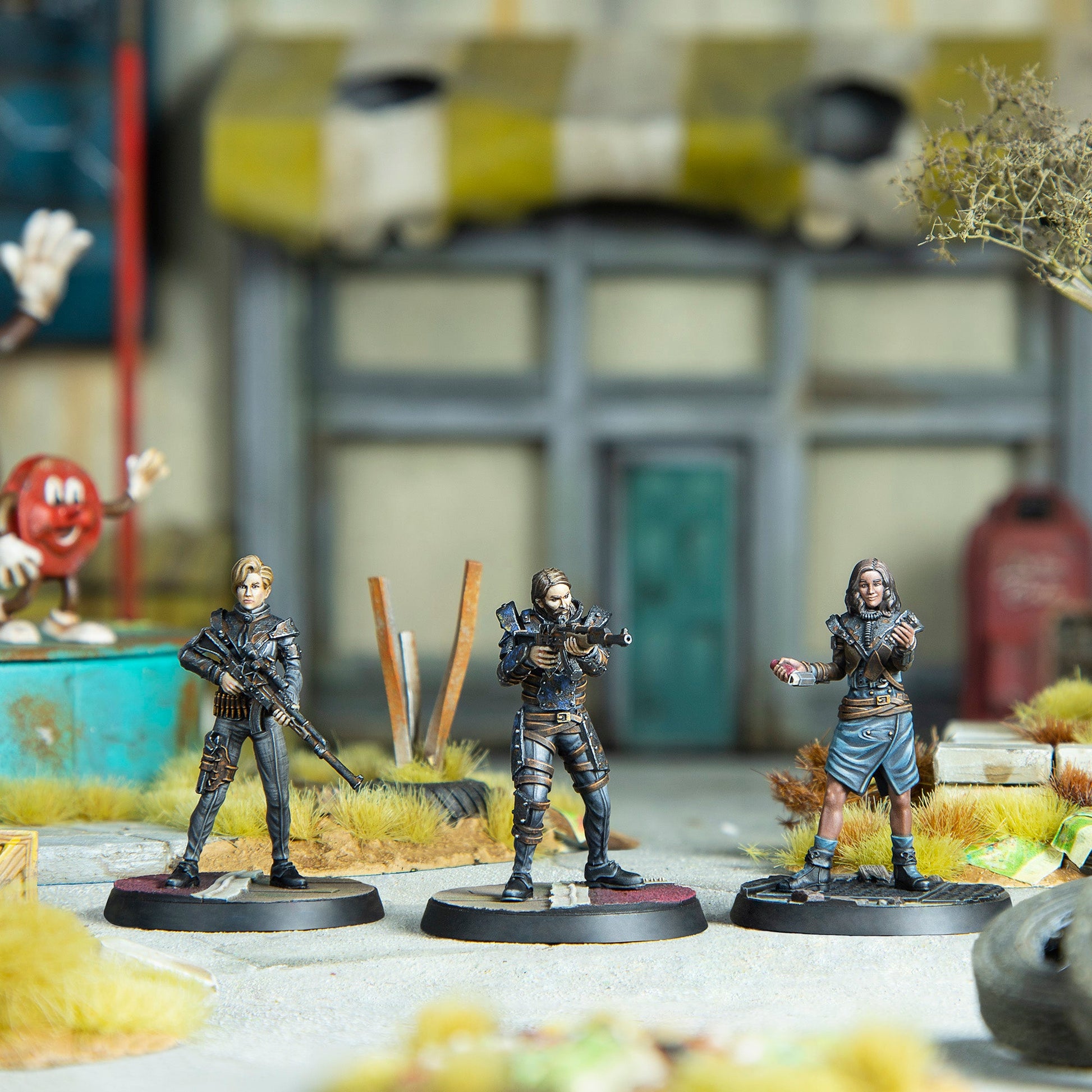 Three miniature figures in a post-apocalyptic diorama - two are armed with rifles; a third holds a book and small object