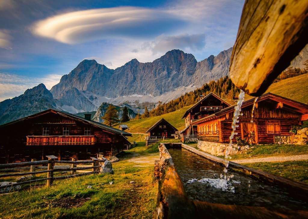 Water flows from a wooden spout into a channel, past rustic alpine chalets nestled on a hillside with towering mountains and a lenticular cloud
