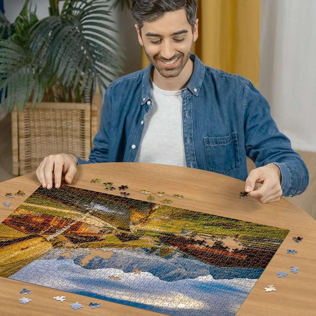 Smiling man in denim shirt assembling a landscape jigsaw puzzle on a wooden table
