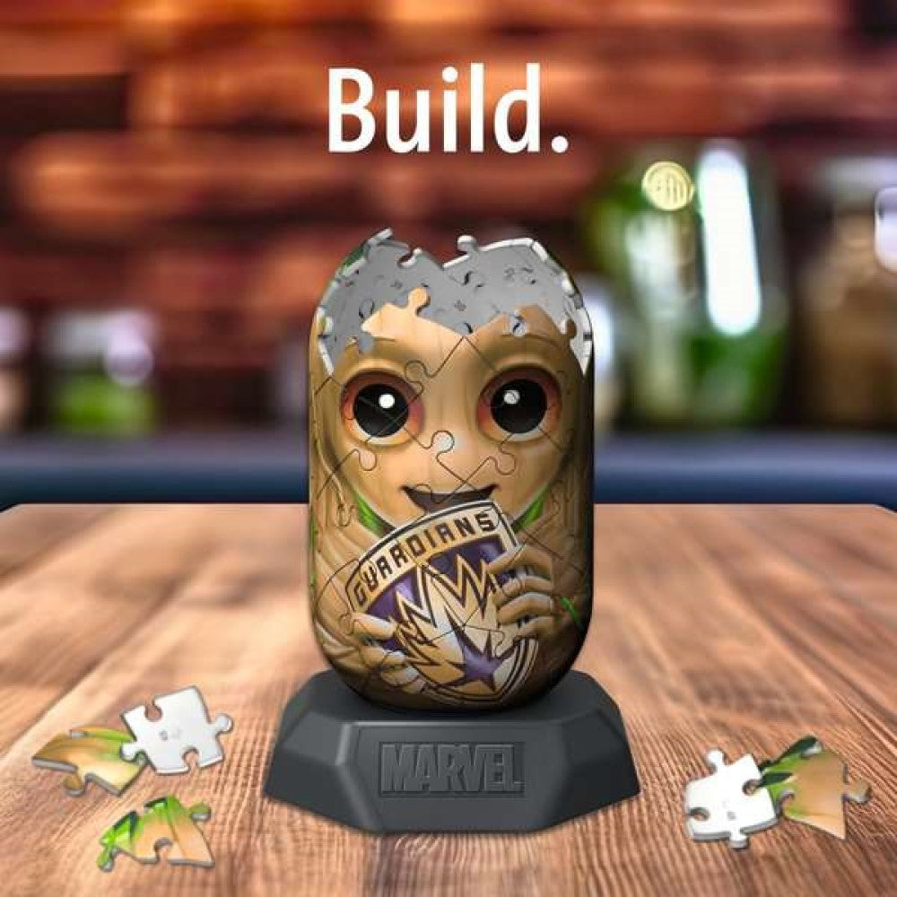 Partially built 3D Baby Groot puzzle on a Marvel stand with loose pieces on a wood table. "Build." text