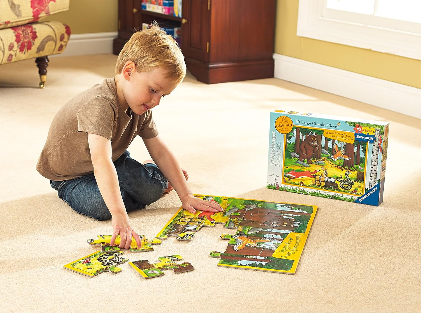 A young blonde boy intently assembles a colorful Gruffalo floor puzzle on a light carpet, with the puzzle box nearby