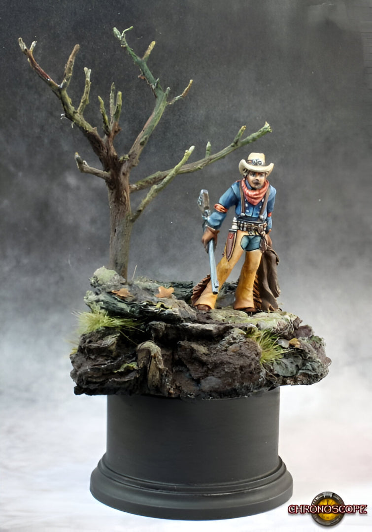 Painted miniature of a cowboy in blue shirt, tan chaps, and hat, holding a rifle, standing on a rocky base next to a bare tree