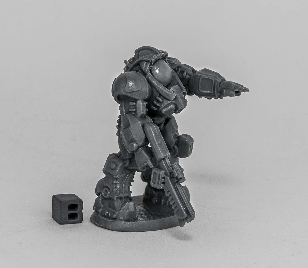 Unpainted grey miniature mech aims with one arm, holds a rifle. Small black 'B' cube provides scale