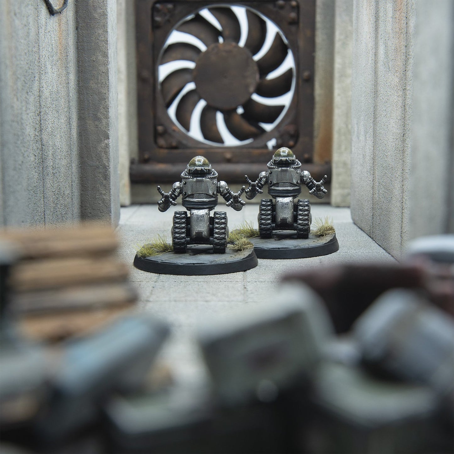 Two silver miniature robots with tank treads and multi-fingered hands stand in a concrete corridor, arms raised, before a large industrial fan.