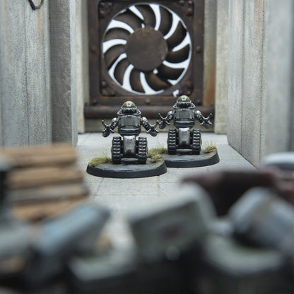Two silver miniature robots with tank treads and multi-fingered hands stand in a concrete corridor, arms raised, before a large industrial fan.