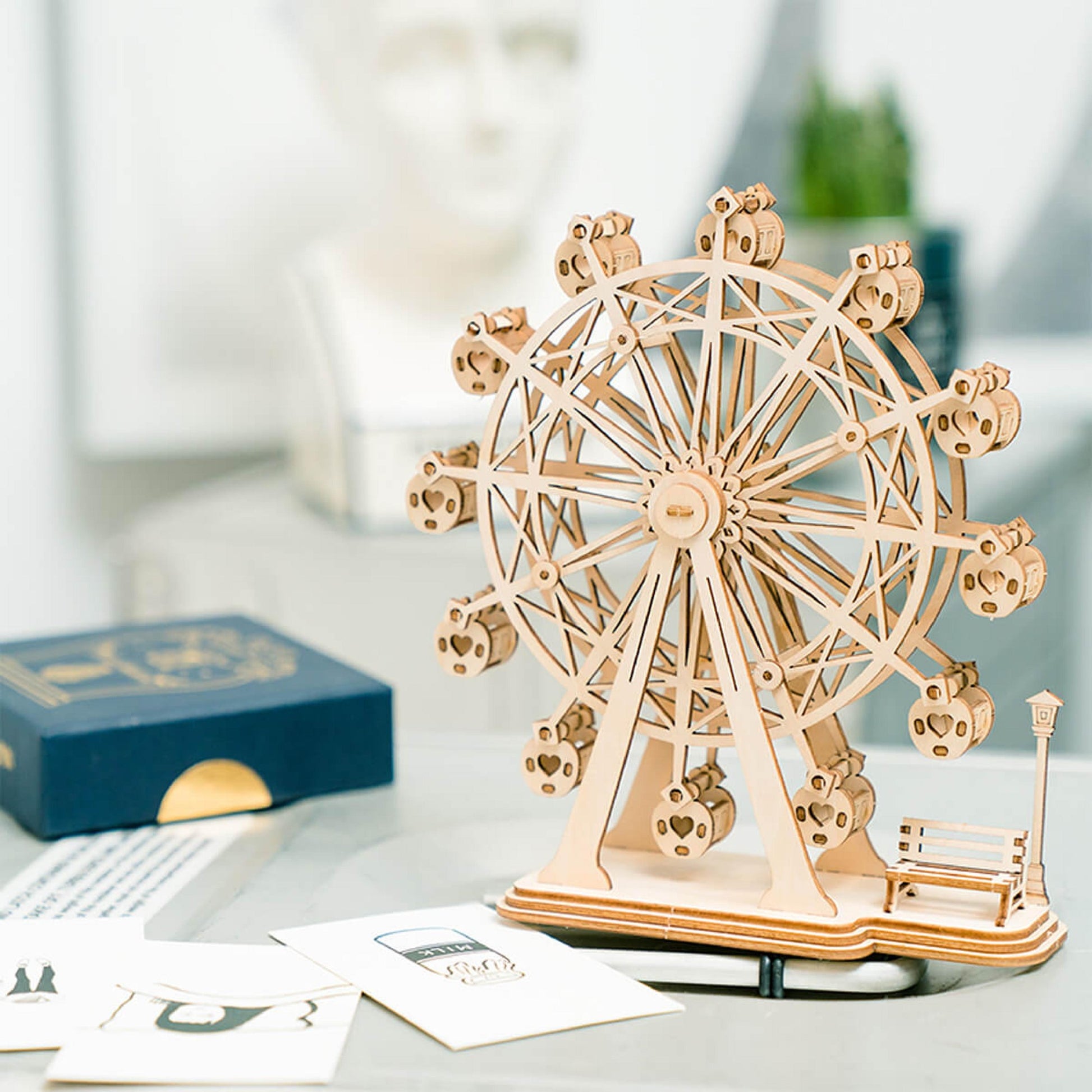 Detailed wooden model of a Ferris wheel, with heart-cutout cabins, displayed on a desk next to cards
