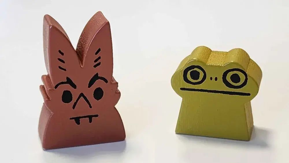Brown bat game piece with fangs, angry face, next to a green frog game piece with big eyes