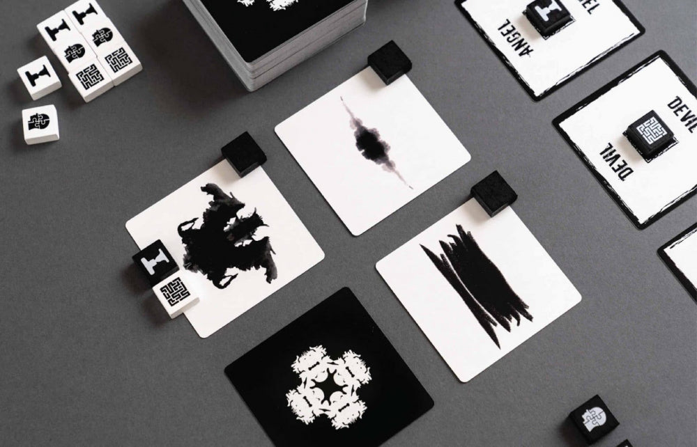 Overhead view of a tabletop game with black and white cards and small dice featuring inkblot designs and stylized symbols