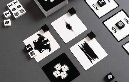 Overhead view of a tabletop game with black and white cards and small dice featuring inkblot designs and stylized symbols