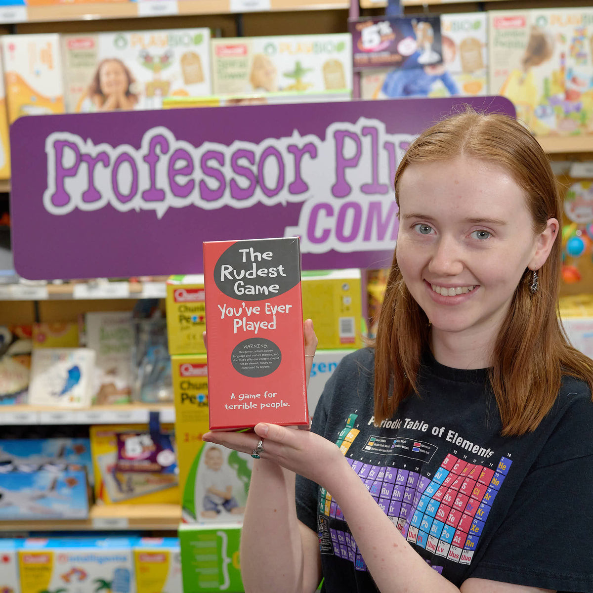 Red-haired woman smiles, holding &quot;The Rudest Game You&#39;ve Ever Played&quot; board game in a store