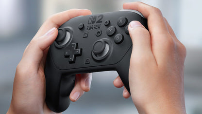 Hands hold a black video game controller featuring a "Nintendo Switch 2" logo and classic button layout