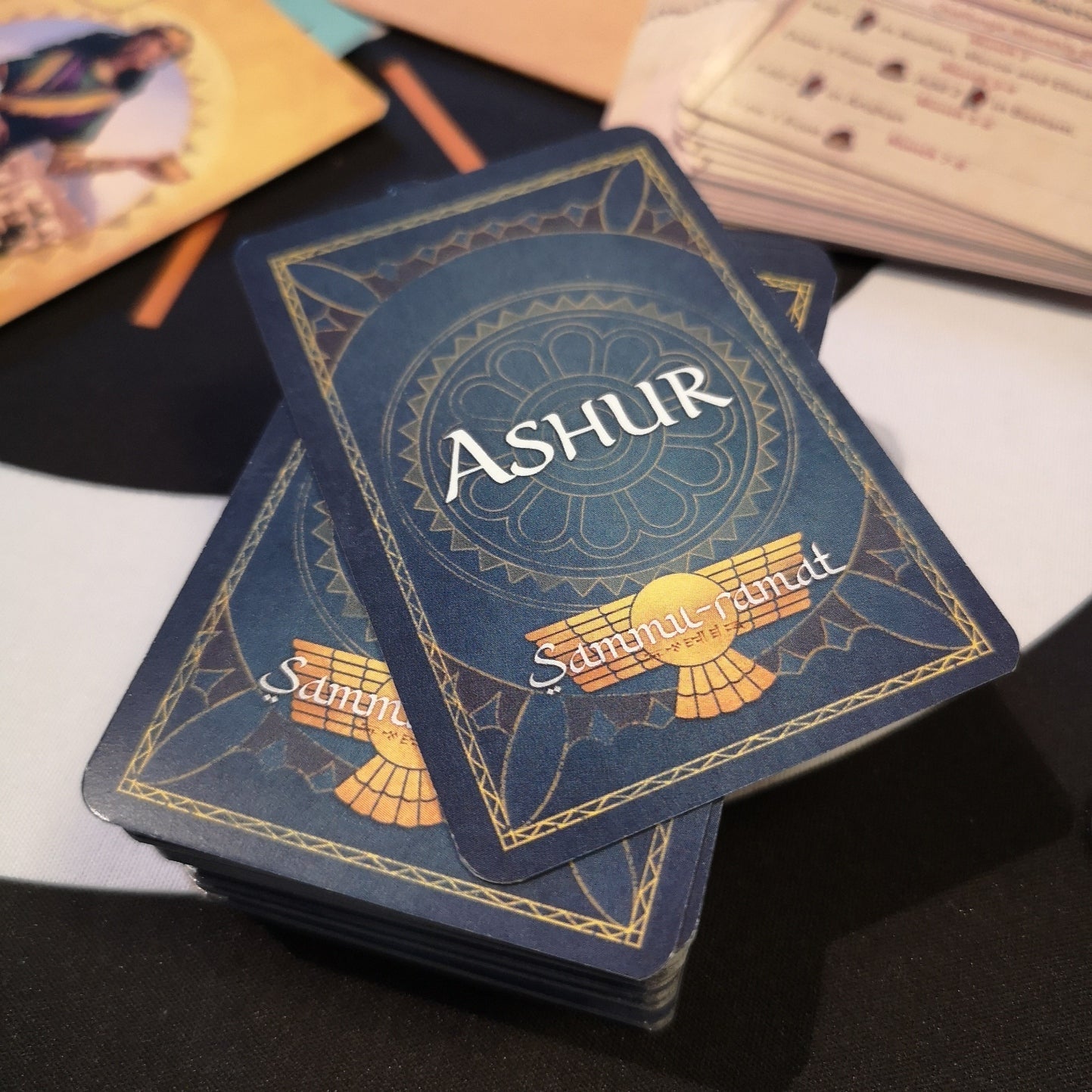Stack of dark blue board game cards top card reads ASHUR Sammul-ramat with a winged sun symbol