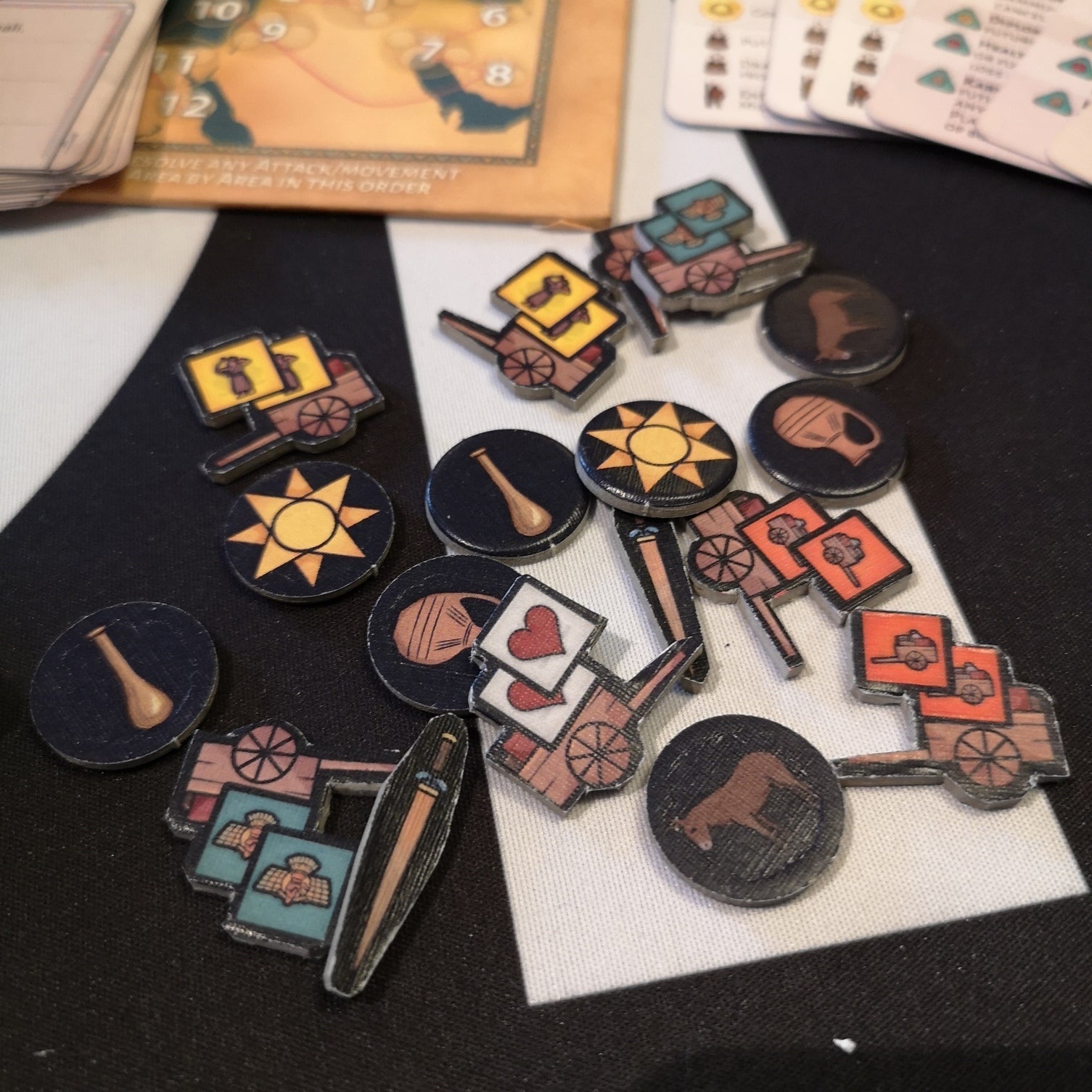 A collection of game pieces including wooden circles with images of a sun pot club and bull and flat wooden pieces depicting carts with different cargo