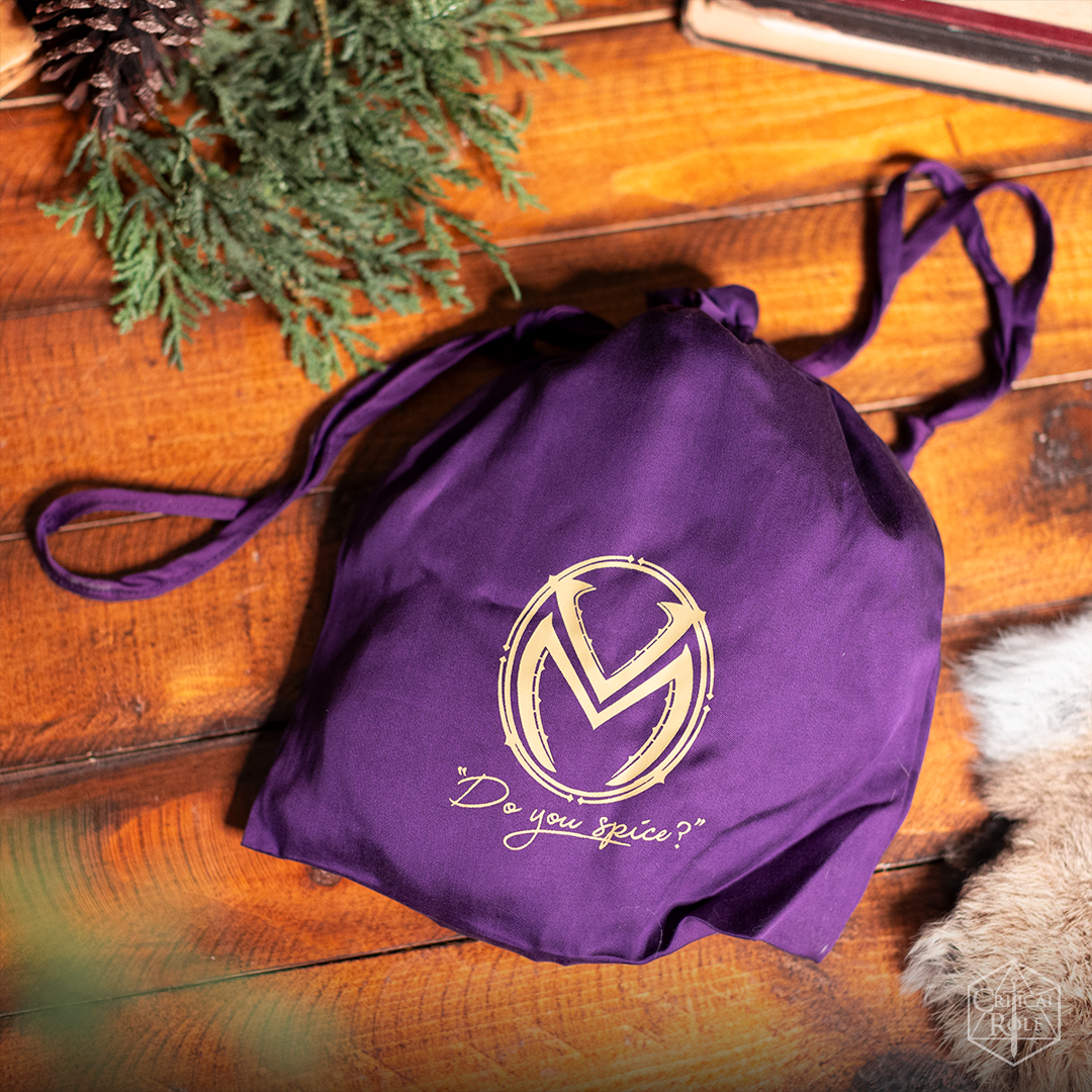 A purple drawstring bag with a gold logo and "Do you spice?" text, on wood with pine boughs, books, and fur