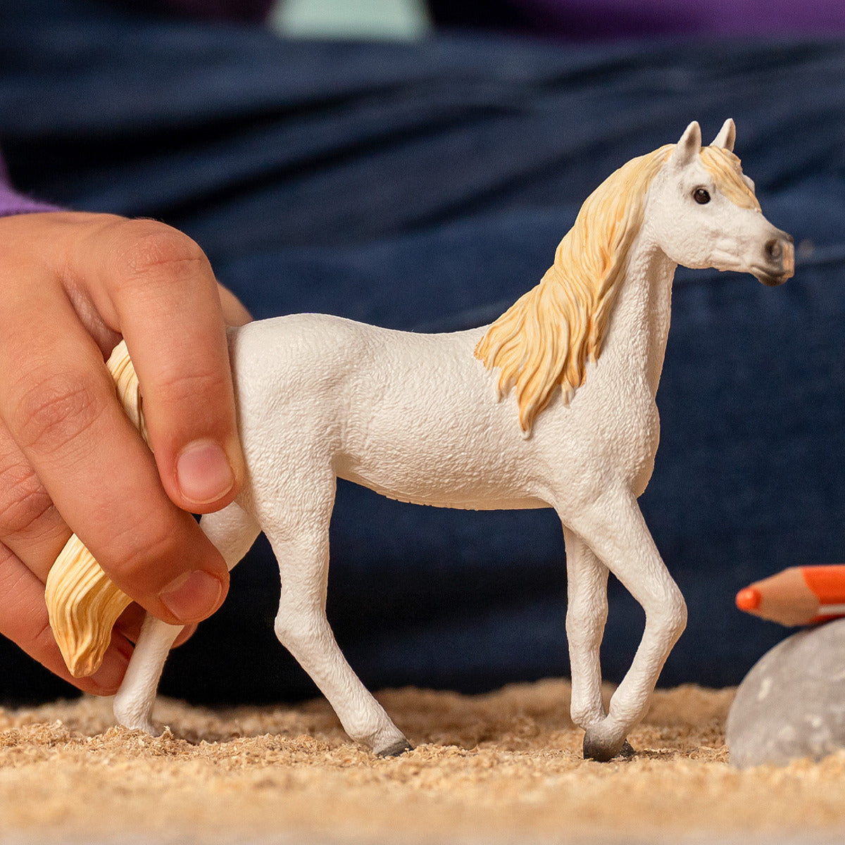 A child&#39;s hand plays with a white Arab Mare toy figurine with a blonde mane on a surface covered in wood shavings